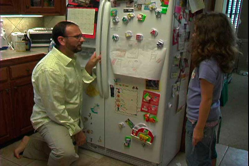 Father kneels next to a refrigerator in the kitchen while speaking to his daughter