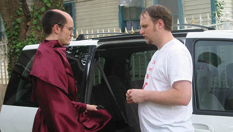 Man in religious robe talks to man in t-shirt outside a mini-van